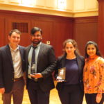 Four people posing for a photo and the two people in the middle are holding a trophy in their hands